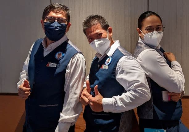 2 male dining stewards and 1 female dining steward pose on Carnival Panorama. Crew are wearing white button down shirts and blue vests and N95 masks.