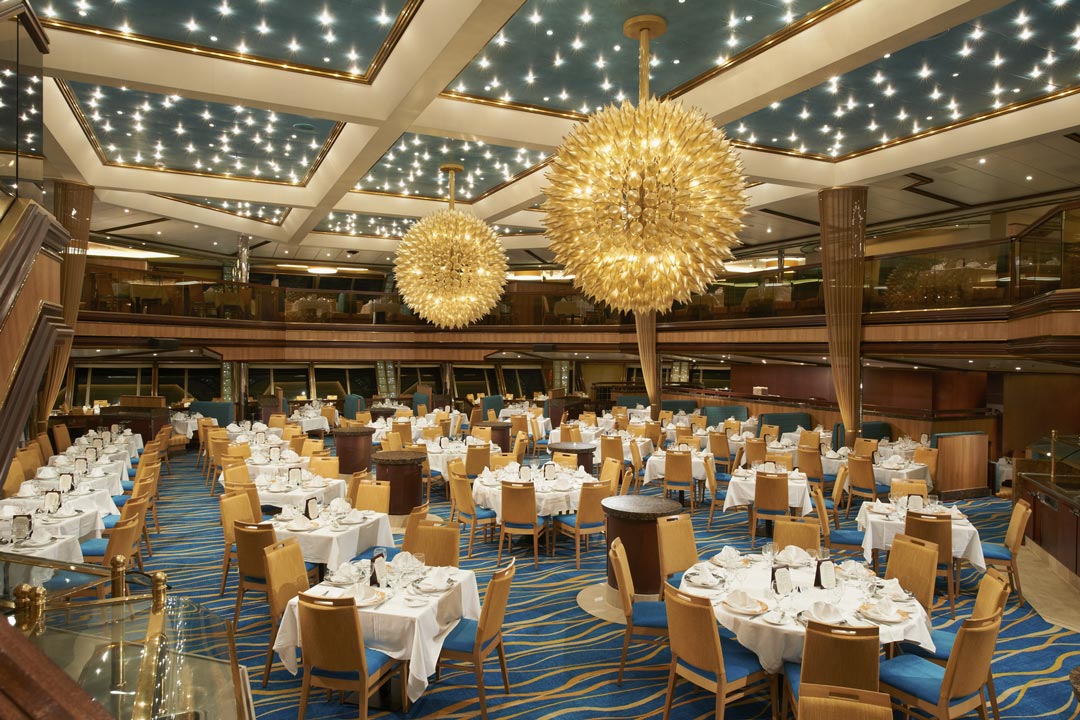 yellow and blue chairs with tables and table cloths and blue and gold carpet in huge dining area with windows in the back and two large chandaleres hang from ceiling