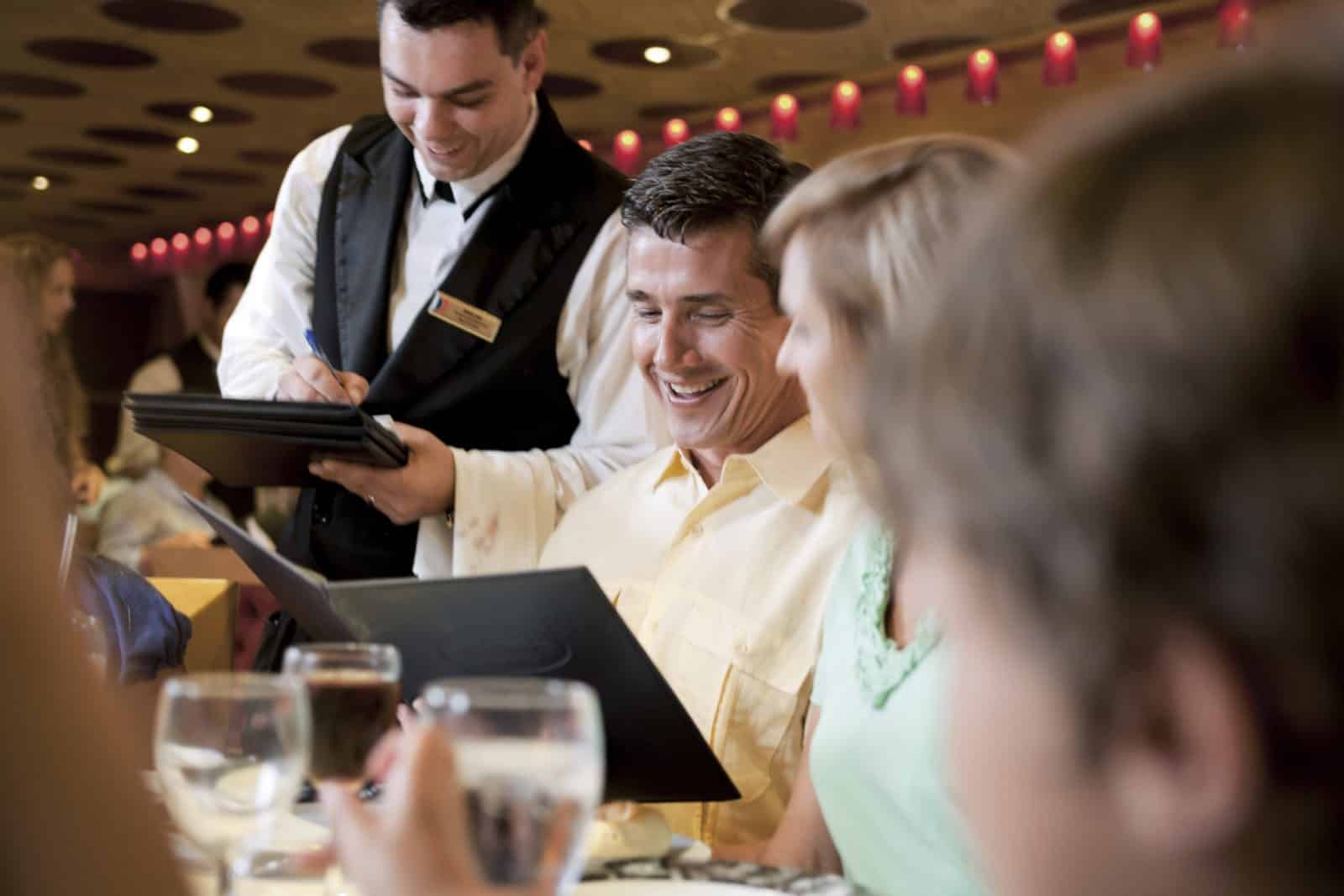 ALT Description: A friendly cruise waiter in a crisp uniform stands attentively beside a well-set table on a luxurious cruise ship. The table is adorned with elegant dinnerware and sparkling glassware, ready to cater to the guests' dining needs. The waiter wears a warm smile, exuding professionalism and hospitality. In the background, the vast expanse of the ocean stretches out, reflecting the serene ambiance of the cruise experience. This image captures the essence of cruising as a premier mode of travel, highlighting the exceptional service, exquisite dining, and breathtaking views that make it a truly unforgettable journey.