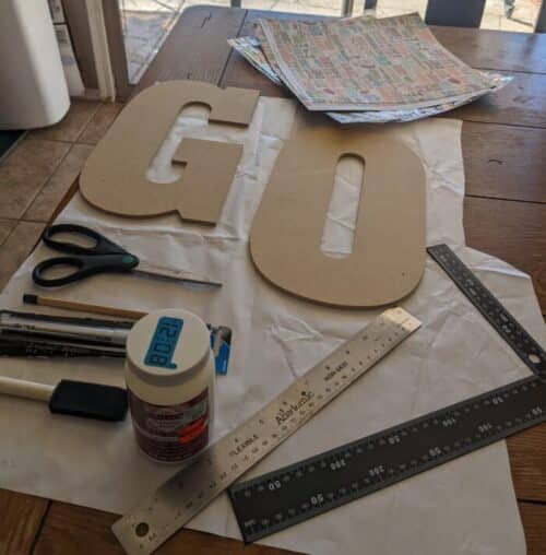 Wooden Letters G & O, ruler, mid podge podge, foam brush and scrapbook paper sit on paper on a table
