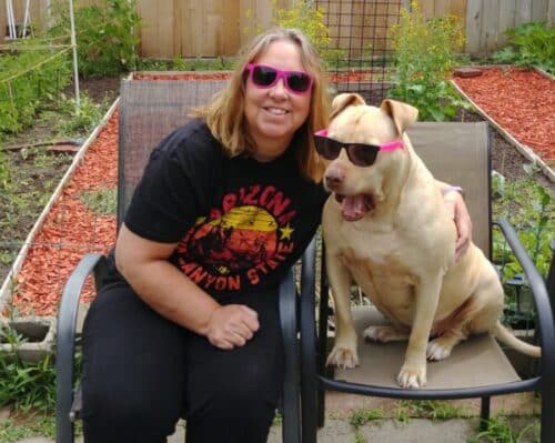 woman and dog pose in sunglasses sitting in chair in front of a vegetable garden