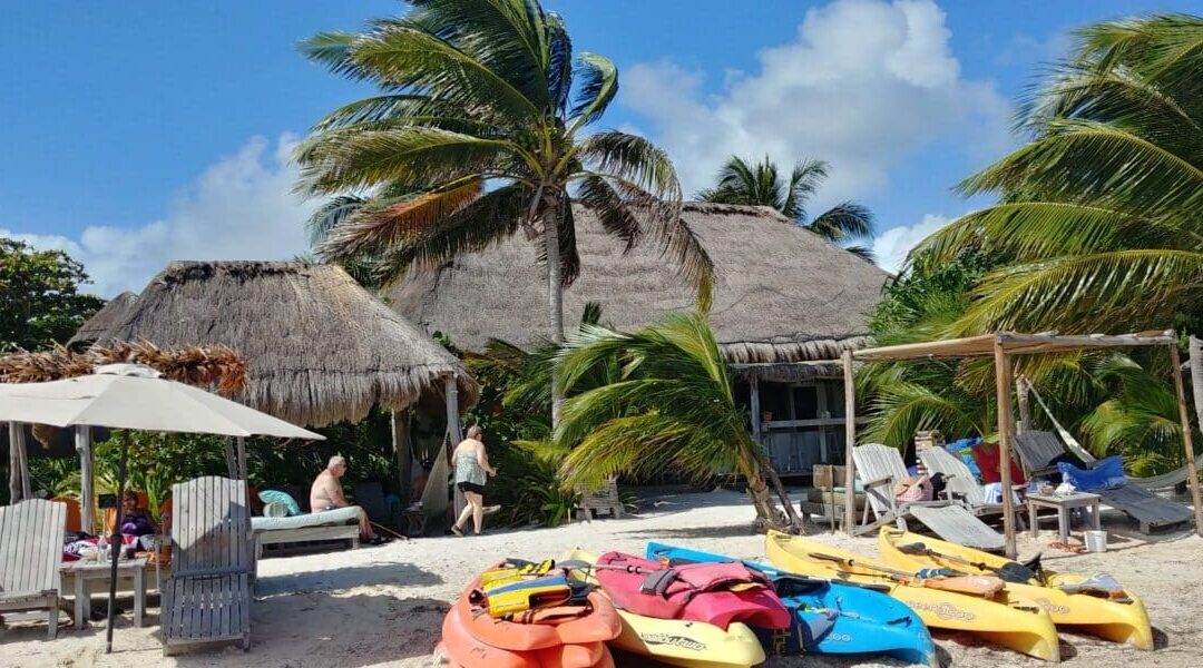 Colorful kayaks and beach toys scattered along the sandy beach, with thatched roof palapas in the background. Sun loungers placed strategically for ultimate relaxation. Pin this image to remember this oasis!