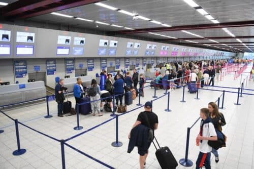 Man in dark colored shirt and shorts pulling roller bag, woman in red and black sweatpants, with white Tshirt waiting-in-line-at-airport-check-in