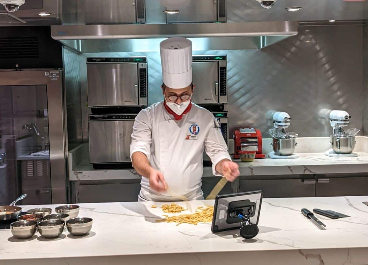 Chef of Carnival Panorama demonstrates how to make pasta at Carnival Kitchen with silver bowls of ingredients and pasta on a white granate conter top.