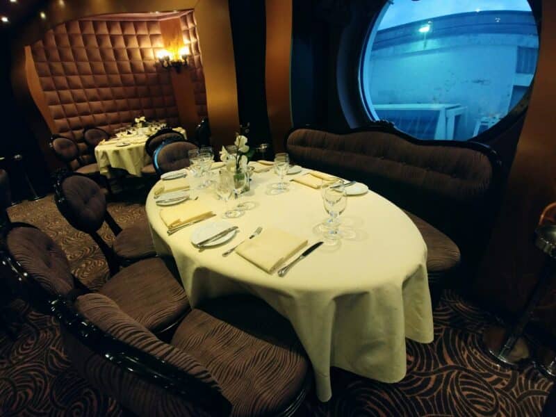 An oval table with a off white table cloth are flanked by dark plush seats. A round window can be seen on MSC Meraviglia.