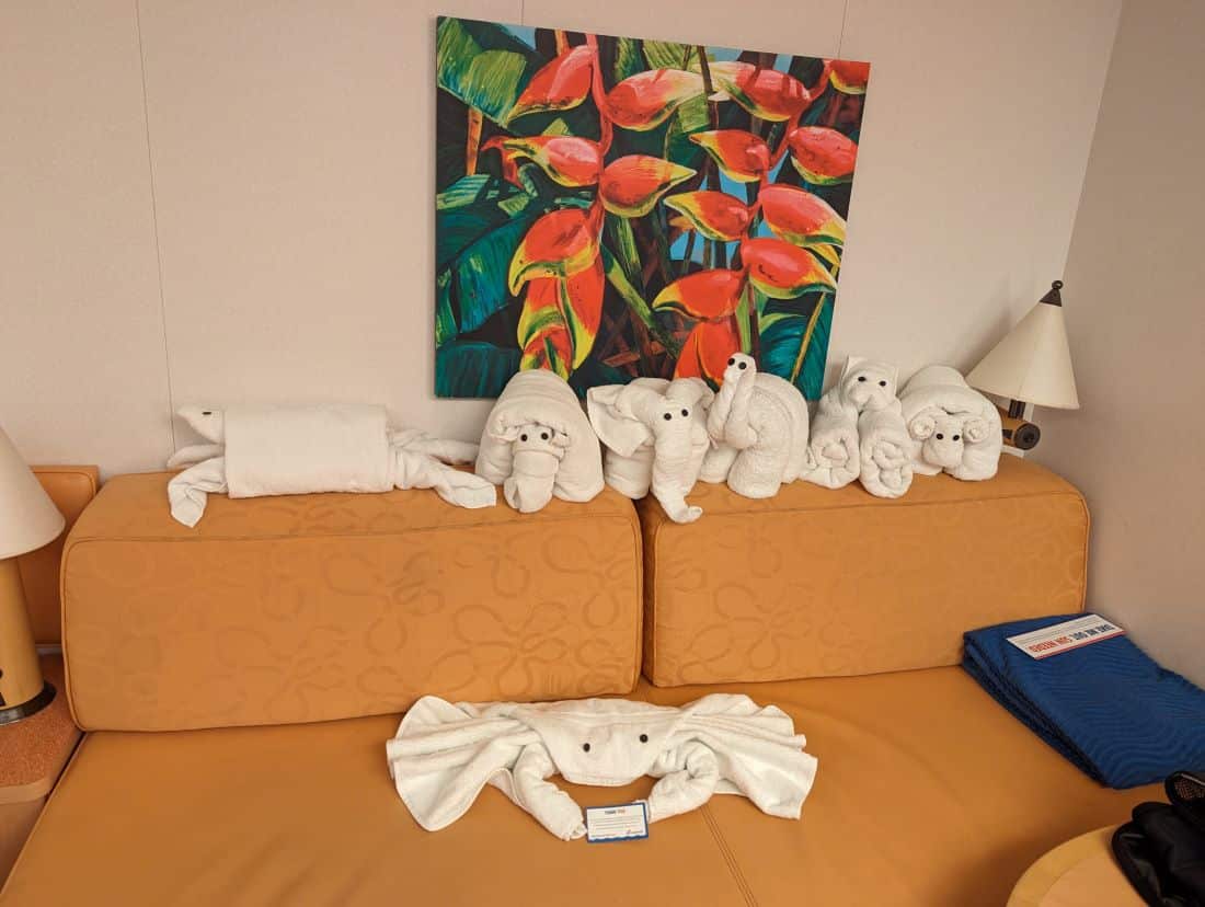 A mustard yellow sofa with several white towel animals and a colorful painting above on Carnival Magic.