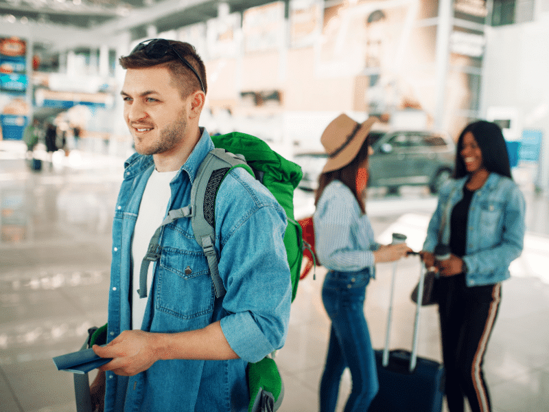 man in blue shirt walks with his passport past two women with suitcases