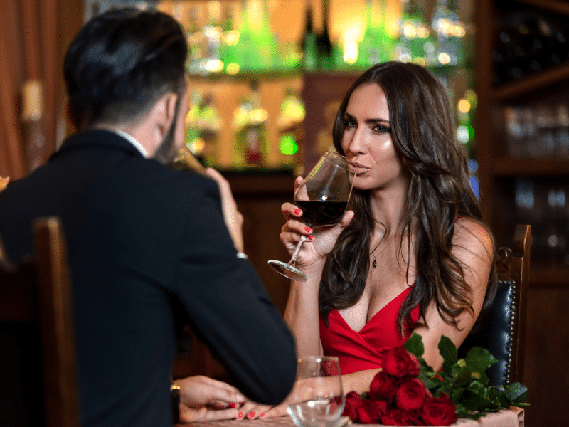 A woman in a red dress sips wine with a man in a black suit coat at a romantic dinner on a Carnival Cruise main dining room