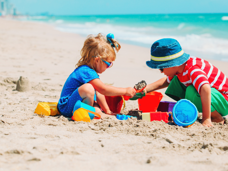 young children play in sand at beach with sun protection of hats, sunglasses, and sun shirts.