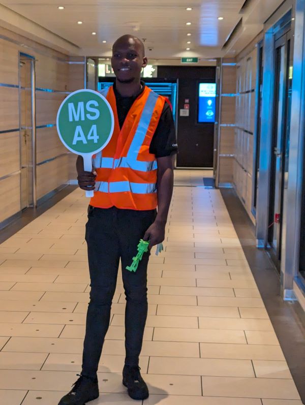 A cruise employee stands with a sign showing which muster station meets in this area.