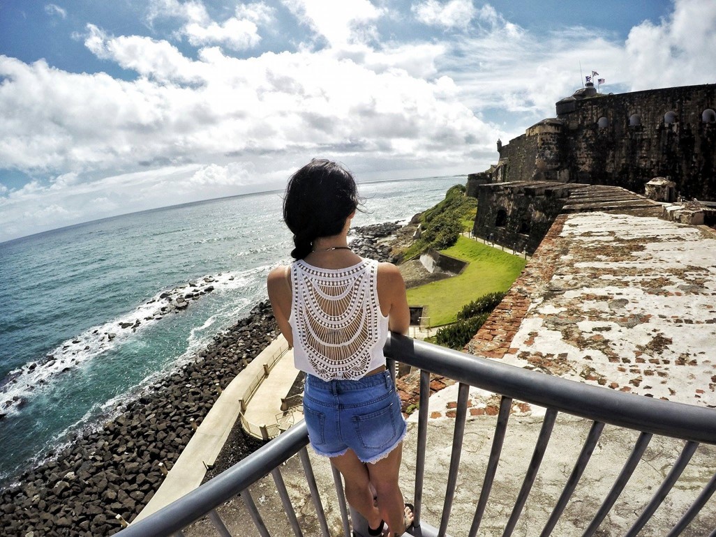 A young woman in a white crocheted top looks out onto the water at an overlook at one of the forts in San Juan